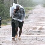 Man covered in a plastic sheet walking through heavy rain and strong wind on a muddy road