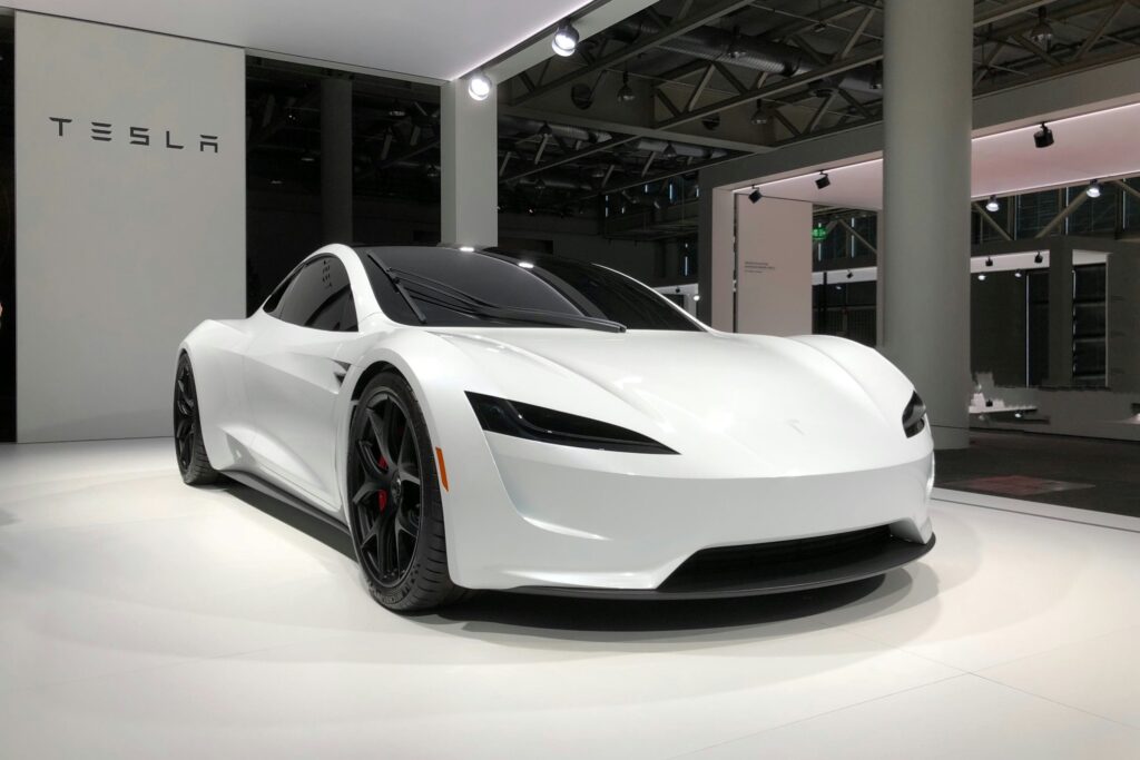 White Tesla Roadster prototype displayed at an exhibit beside a Tesla wall sign